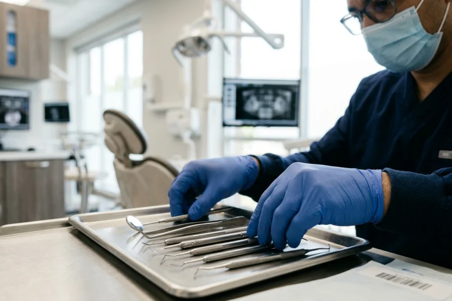 Close de mãos de um dentista profissional organizando instrumentos clínicos em uma bandeja de aço inoxidável em um consultório moderno e iluminado.
